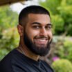 Close up headshot of a Pakistani man looking at the camera while smiling in a garden in summer. He is in the North East of England.